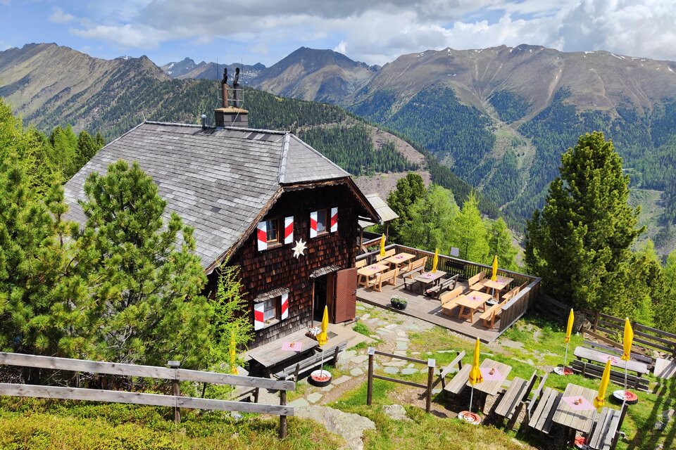 Eine gemütliche Berghütte umgeben von grünen Bäumen und Bergen. Draußen stehen Holztische mit gelben Sonnenschirmen. | © TV Murau