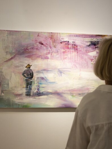 A person stands in front of a modern painting that is done in soft colors. The artwork shows a figurative representation that is surrounded by an abstract landscape. | © Achromatic Photography