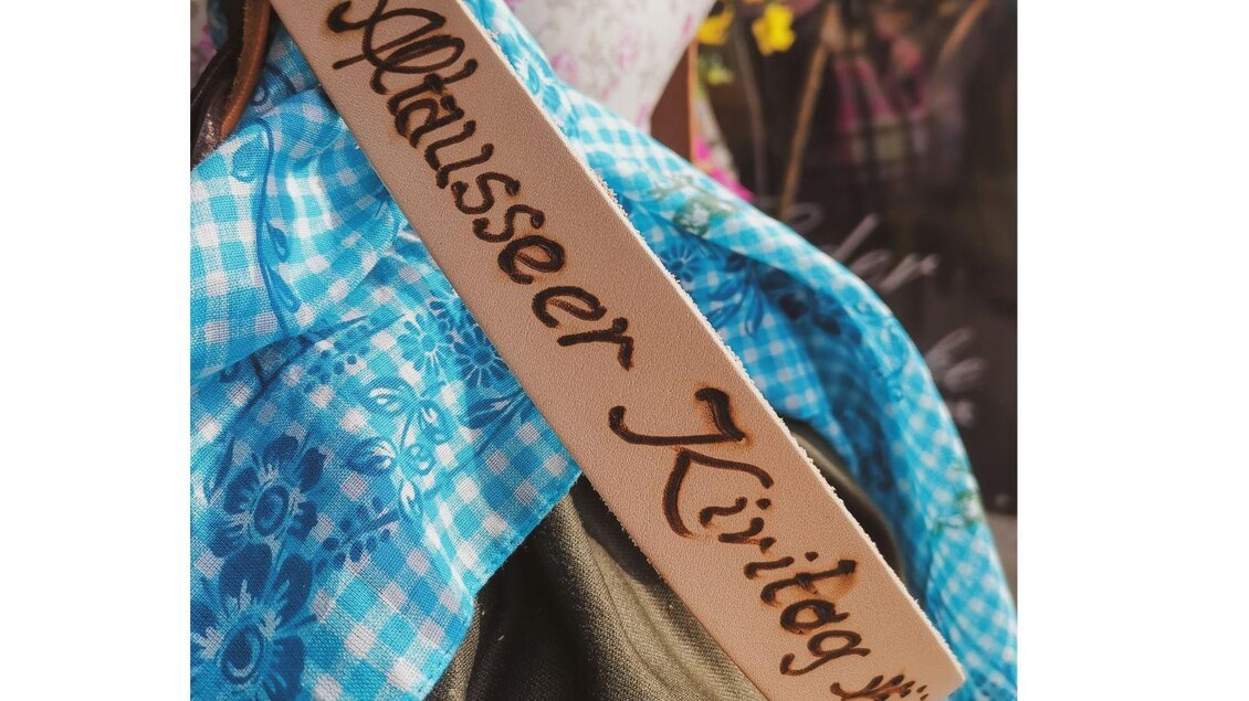 Ein traditionelles Kleidungsstück mit einer blauen karierten Bluse und einer Lederschärpe. Auf der Schärpe steht "Ausseer Kirttag" in kunstvoller Schrift. | © TVB Ausseerland Salzkammergut_Theresa Schwaiger
