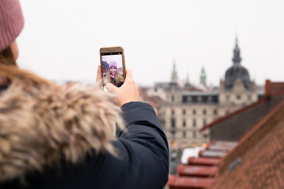 Eine Person macht ein Selfie mit einem Smartphone, während sie auf eine Stadtlandschaft blickt. Im Hintergrund sind historische Gebäude und eine graue Himmel zu sehen. | © Graz Tourismus - Katja Stadler