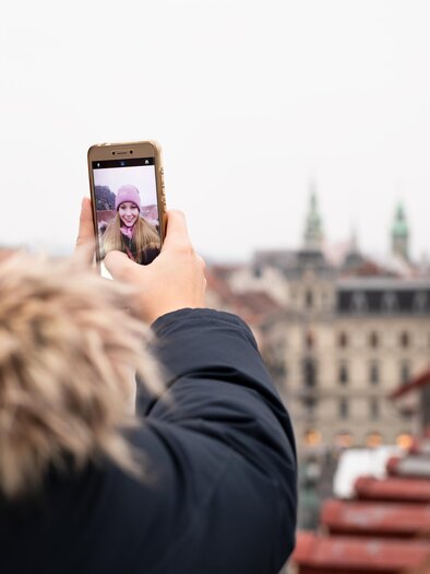 A person is taking a selfie with a smartphone while looking at a cityscape. In the background, historical buildings and a gray sky can be seen. | © Graz Tourismus - Katja Stadler