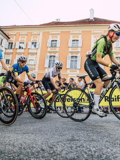 A group of cyclists is participating in a race. In the background, yellow signs and a building are visible. | © david pichler