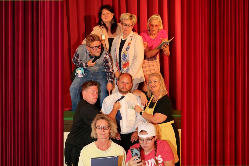 Eine Gruppe von acht Personen posiert auf einer Bühne vor einem roten Vorhang. Die Stimmung ist humorvoll und lebhaft, während sie unterschiedliche Posen und Gesichtsausdrücke zeigen. | © Theatergruppe Deutsch Goritz