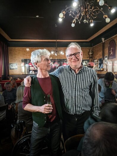 Two men are standing in a cozy bar, smiling at each other. In the background, there are other guests and atmospheric lighting to be seen. | © Holzmann