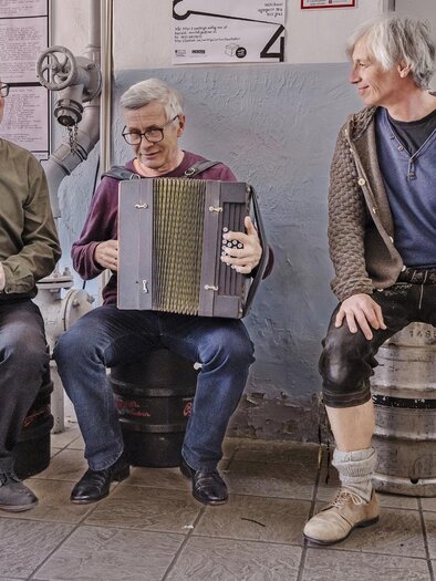 Three men are sitting on barrels and playing music together. One is playing the flute, one an accordion, and the third a violin. | © Kranzlbinder