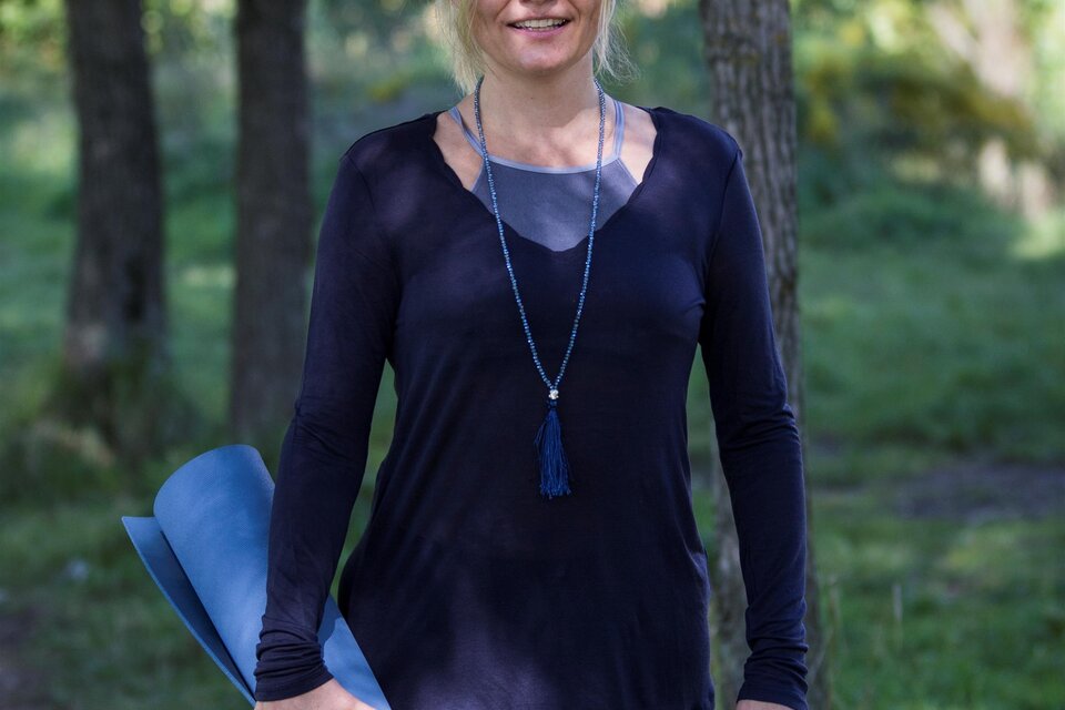 Eine Frau steht in einem Wald und hält eine blaue Yogamatte. Sie trägt ein schwarzes Oberteil und schaut freundlich in die Kamera. | © Anjali Yoga