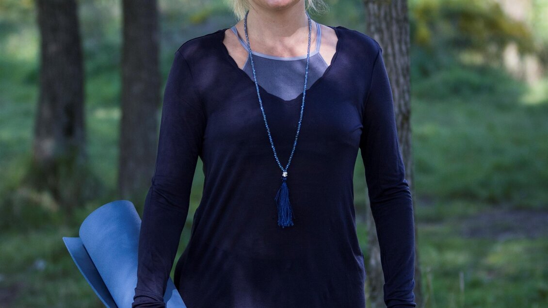 Eine Frau steht in einem Wald und hält eine blaue Yogamatte. Sie trägt ein schwarzes Oberteil und schaut freundlich in die Kamera. | © Anjali Yoga