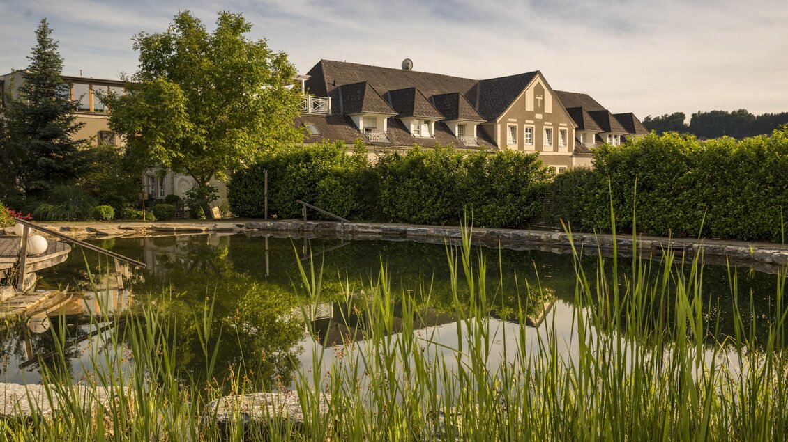 Ein ruhiger Teich spiegelt ein Gebäude und die umgebende Natur wider. Die Umgebung ist grün und einladend. | © Garten Hotel Ochensberger
