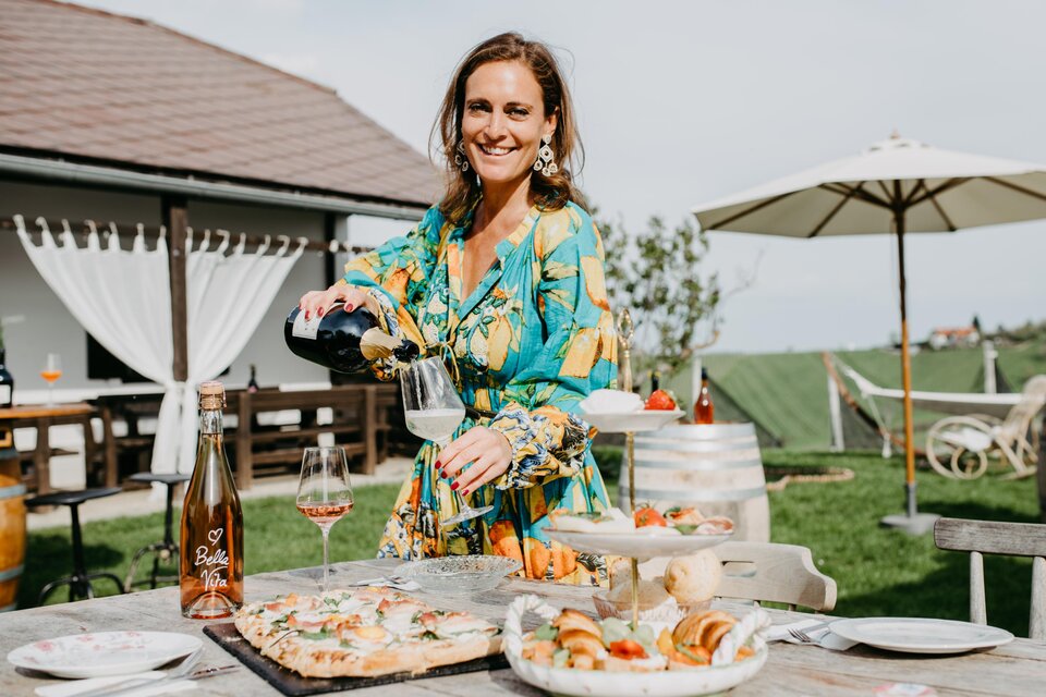 Eine fröhliche Frau serviert Getränke und Essen auf einem Tisch im Freien. Im Hintergrund sind ein sonniger Garten und ein überdachter Sitzbereich zu sehen. | © Conny Leitgeb