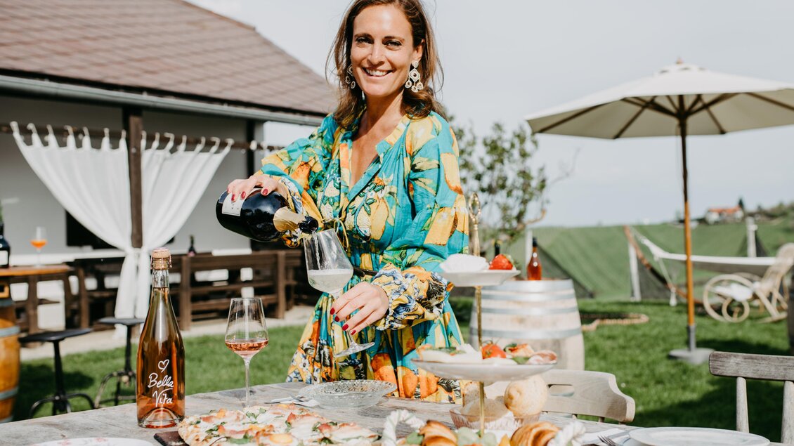 Eine fröhliche Frau serviert Getränke und Essen auf einem Tisch im Freien. Im Hintergrund sind ein sonniger Garten und ein überdachter Sitzbereich zu sehen. | © Conny Leitgeb
