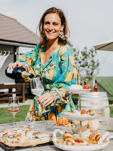 Eine fröhliche Frau serviert Getränke und Essen auf einem Tisch im Freien. Im Hintergrund sind ein sonniger Garten und ein überdachter Sitzbereich zu sehen. | © Conny Leitgeb