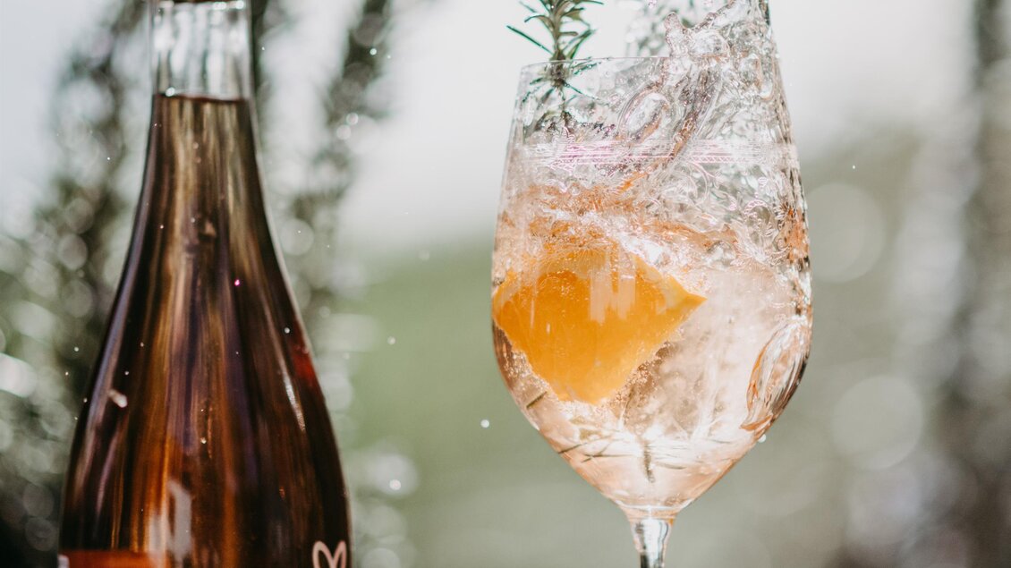 Ein erfrischendes Getränk in einem eleganten Glas sprudelt, umgeben von einer Flasche Sekt und einer halbierten Zitrone. Im Hintergrund sind grüne Pflanzen sichtbar. | © Conny Leitgeb