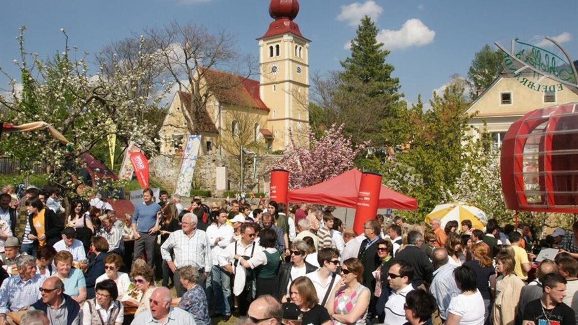 Ein lebhaftes Fest mit vielen Menschen, die zusammen feiern. Im Hintergrund sieht man eine historische Kirche und blühende Bäume. | © Tourismusverband Oststeiermark/Walter Schneider
