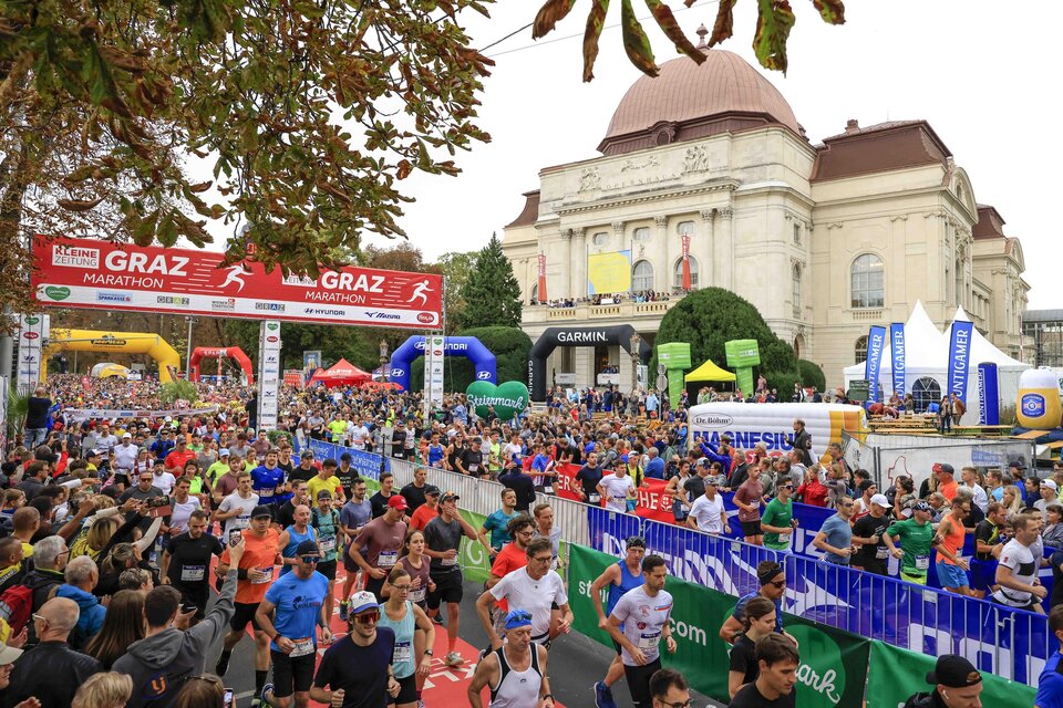 Eine große Menschenmenge läuft beim Graz Marathon. Im Hintergrund sind bunten Banner und ein historisches Gebäude sichtbar.
