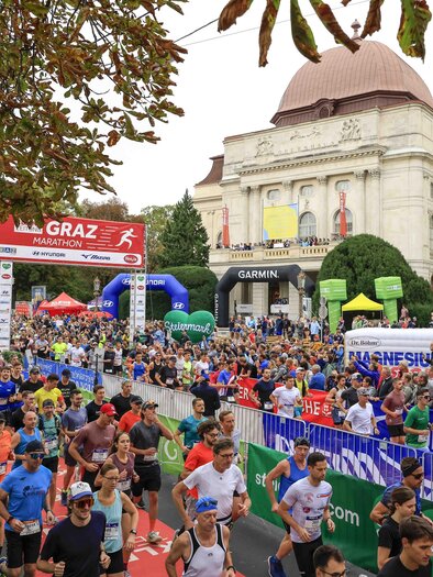A large crowd is running at the Graz Marathon. Colorful banners and a historical building are visible in the background.