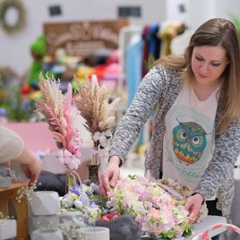 Eine kreative Floristin arrangiert Blumen und Gräser auf einem Tisch. Der Hintergrund zeigt eine bunte Auswahl an Bastelmaterialien. | © Adobe Stock