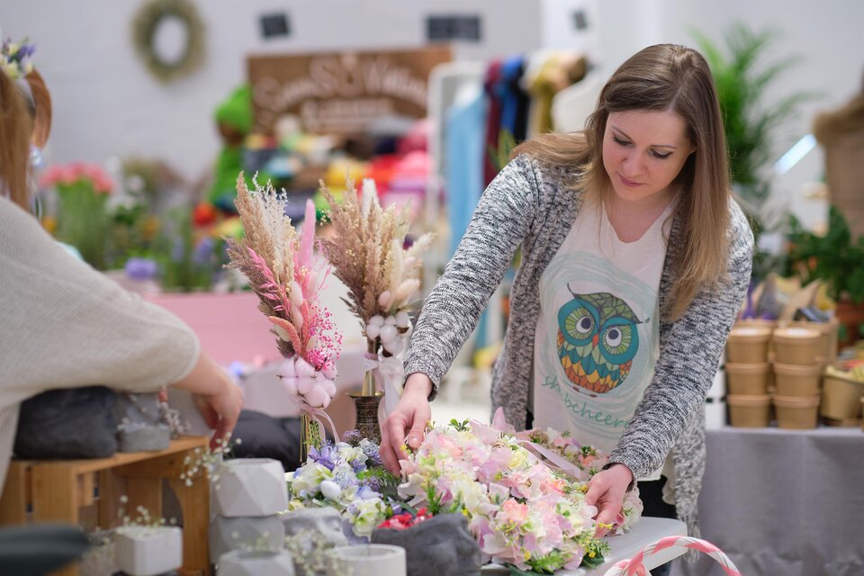 Eine kreative Floristin arrangiert Blumen und Gräser auf einem Tisch. Der Hintergrund zeigt eine bunte Auswahl an Bastelmaterialien. | © Adobe Stock