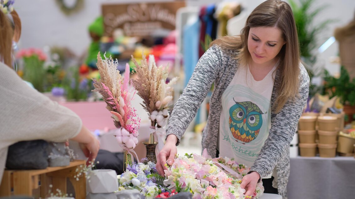 Eine kreative Floristin arrangiert Blumen und Gräser auf einem Tisch. Der Hintergrund zeigt eine bunte Auswahl an Bastelmaterialien. | © Adobe Stock