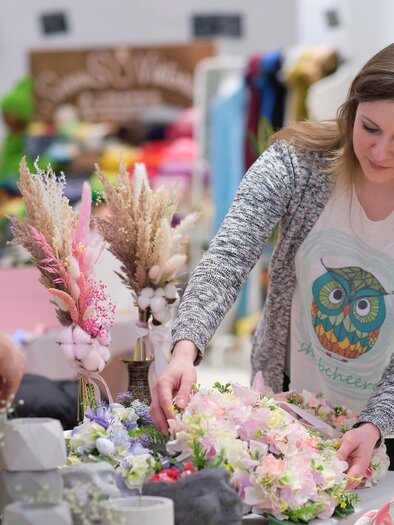 Eine kreative Floristin arrangiert Blumen und Gräser auf einem Tisch. Der Hintergrund zeigt eine bunte Auswahl an Bastelmaterialien. | © Adobe Stock