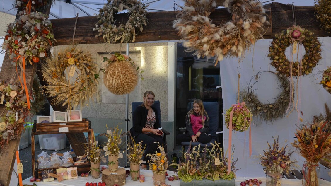 Ein schöner Stand mit herbstlichen Kränzen und dekorativen Arrangements aus getrockneten Blumen und Pflanzen. Zwei Frauen sitzen hinter dem Tisch, der mit verschiedenen handgemachten Produkten geschmückt ist. | © Forum Arnfels
