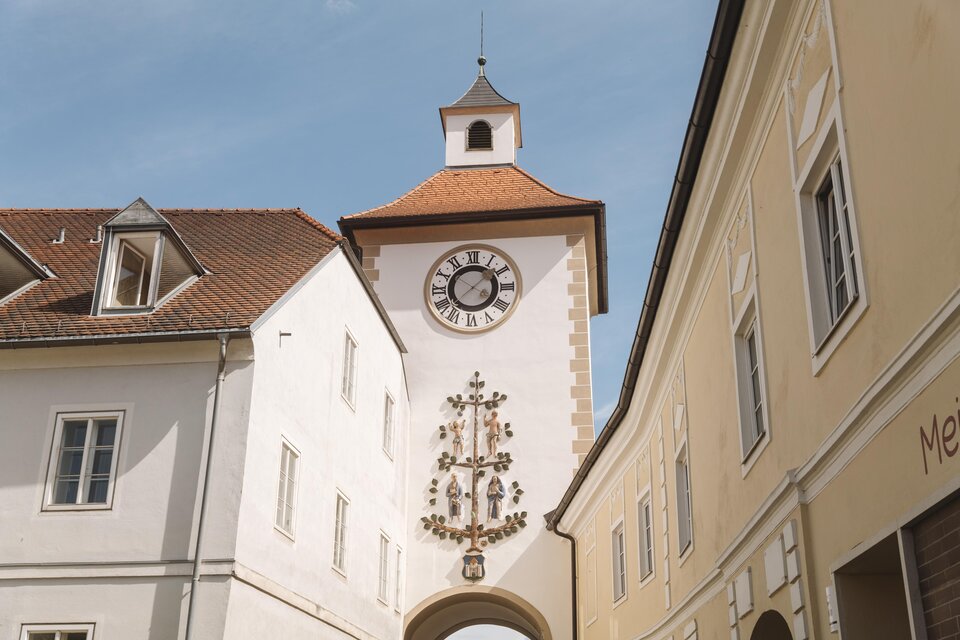 Ein historischer Uhrturm mit einem auffälligen Ziffernblatt und einem roten Dach. Er befindet sich zwischen weißen Gebäuden in einer engen Gasse. | © Karl Steinegger