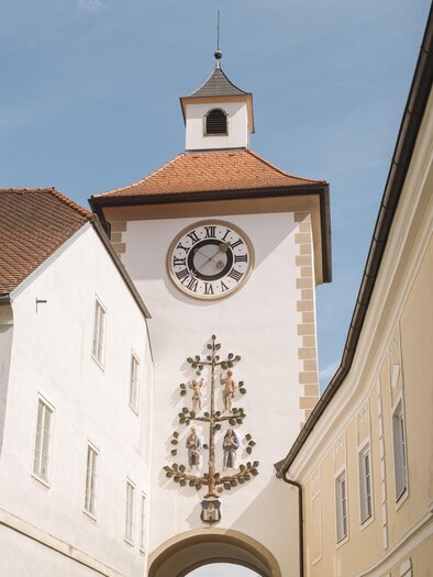 Ein historischer Uhrturm mit einem auffälligen Ziffernblatt und einem roten Dach. Er befindet sich zwischen weißen Gebäuden in einer engen Gasse. | © Karl Steinegger