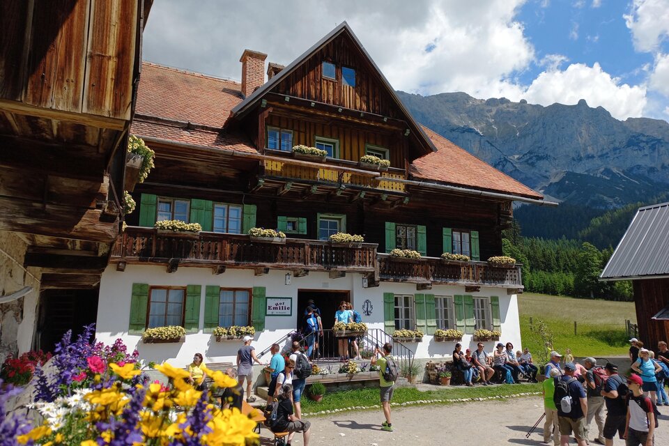 Ein traditionelles, malerisches Almhaus mit Blumenkästen und grünem Holz. Viele Menschen versammeln sich davor, während die Berge im Hintergrund sichtbar sind. | © Tourismusverband Schladming-Dachstein