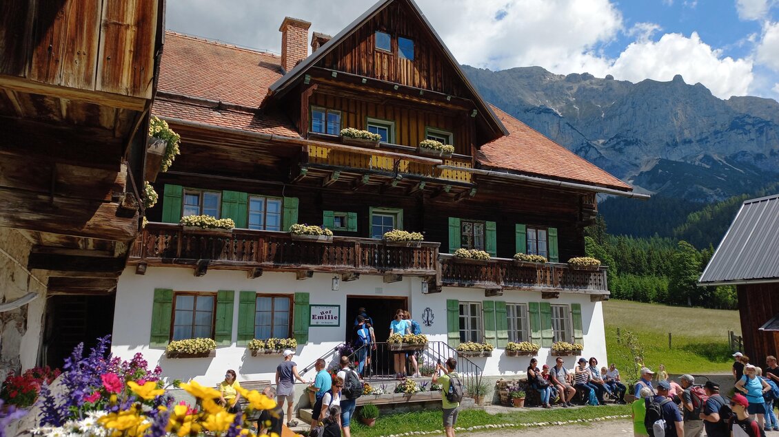 Ein traditionelles, malerisches Almhaus mit Blumenkästen und grünem Holz. Viele Menschen versammeln sich davor, während die Berge im Hintergrund sichtbar sind. | © Tourismusverband Schladming-Dachstein