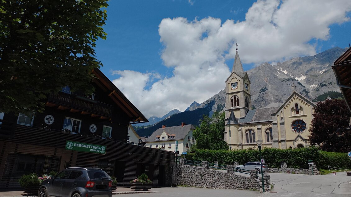 Eine malerische Straße mit traditionellen Holzhäusern und einer beeindruckenden Kirche. Im Hintergrund sind Berge und ein blauer Himmel mit weißen Wolken zu sehen. | © Tourismusverband Schladming-Dachstein
