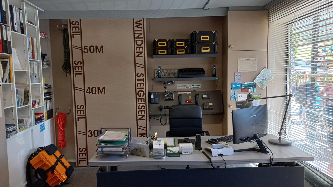 Ein Büro mit einem Schreibtisch, auf dem Computer und Unterlagen liegen. Im Hintergrund sind Regale mit Materialien und Werkzeugen zu sehen. | © Tourismusverband Schladming-Dachstein