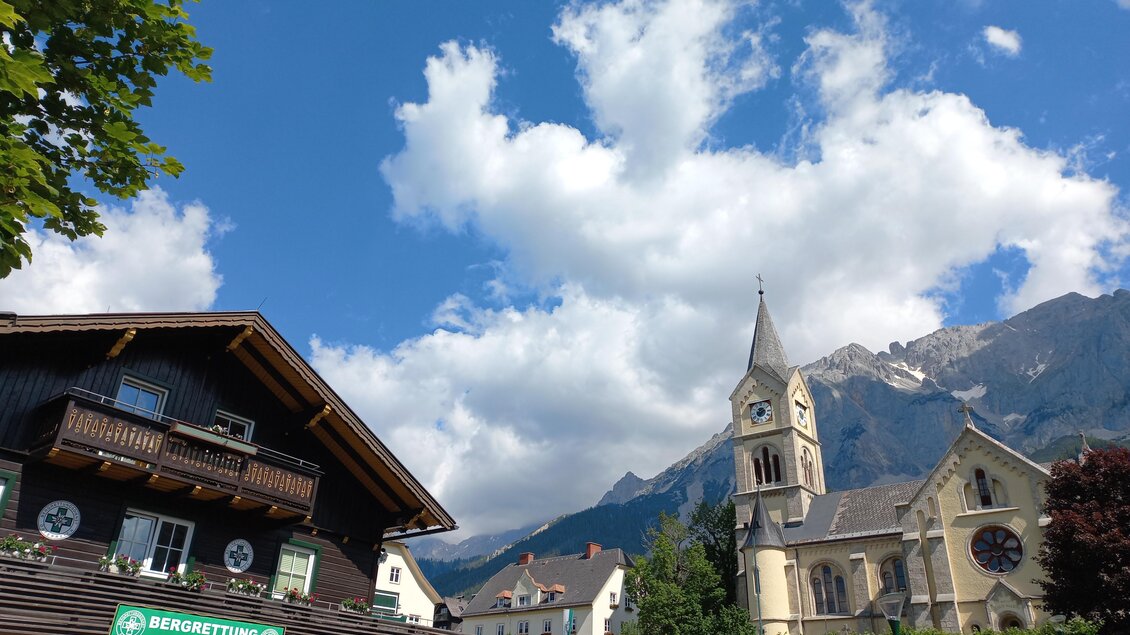 Eine malerische Szene mit einem traditionellen Gebäude und einer Kirche vor einer bergigen Landschaft. Der Himmel ist blau mit vereinzelten Wolken. | © Tourismusverband Schladming-Dachstein