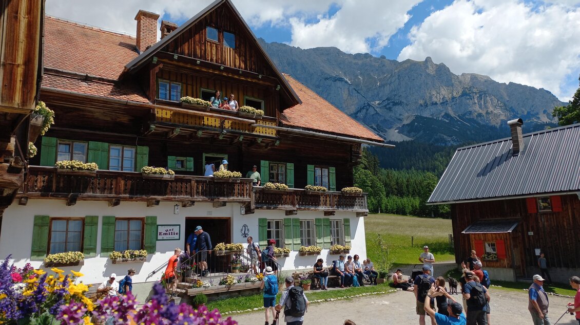 Ein traditionelles altes Bauernhaus mit Balkonen und blühenden Blumen. Menschen versammeln sich im Vordergrund, während im Hintergrund majestätische Berge zu sehen sind. | © Tourismusverband Schladming-Dachstein