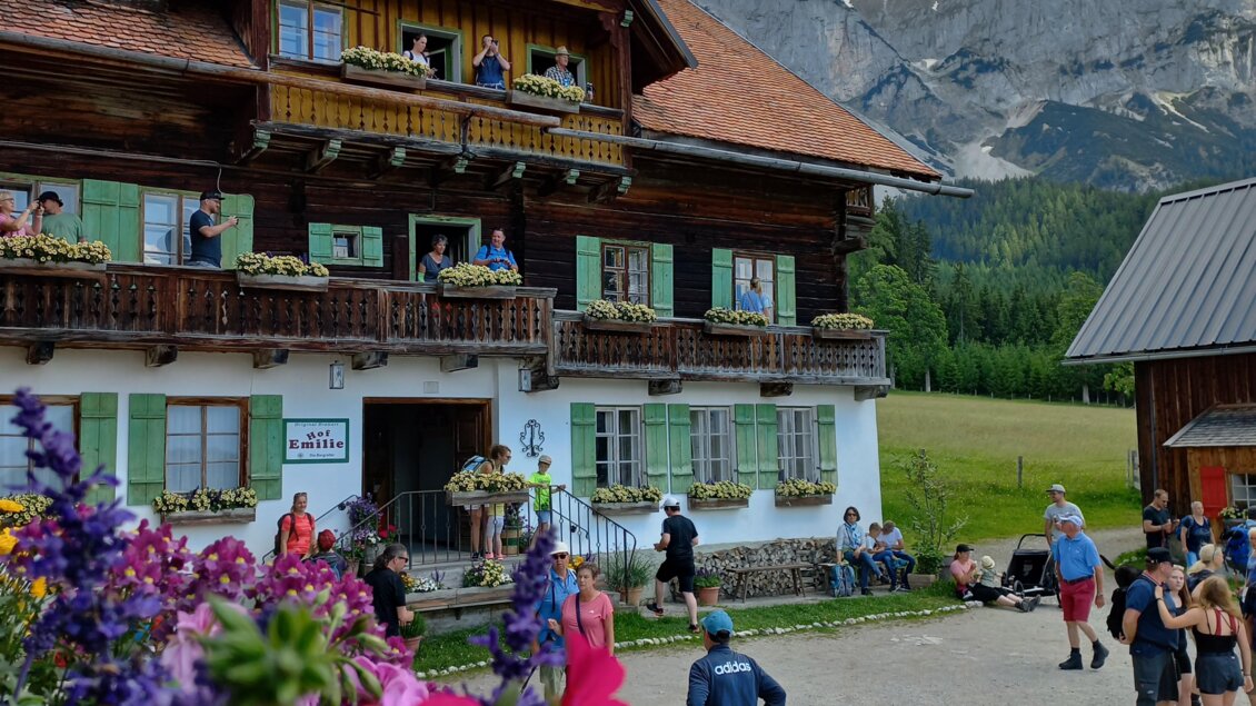 Ein malerisches Holzhaus mit Balkonen, umgeben von bunten Blumen. Im Vordergrund sind Menschen und eine wunderschöne Berglandschaft zu sehen. | © Tourismusverband Schladming-Dachstein