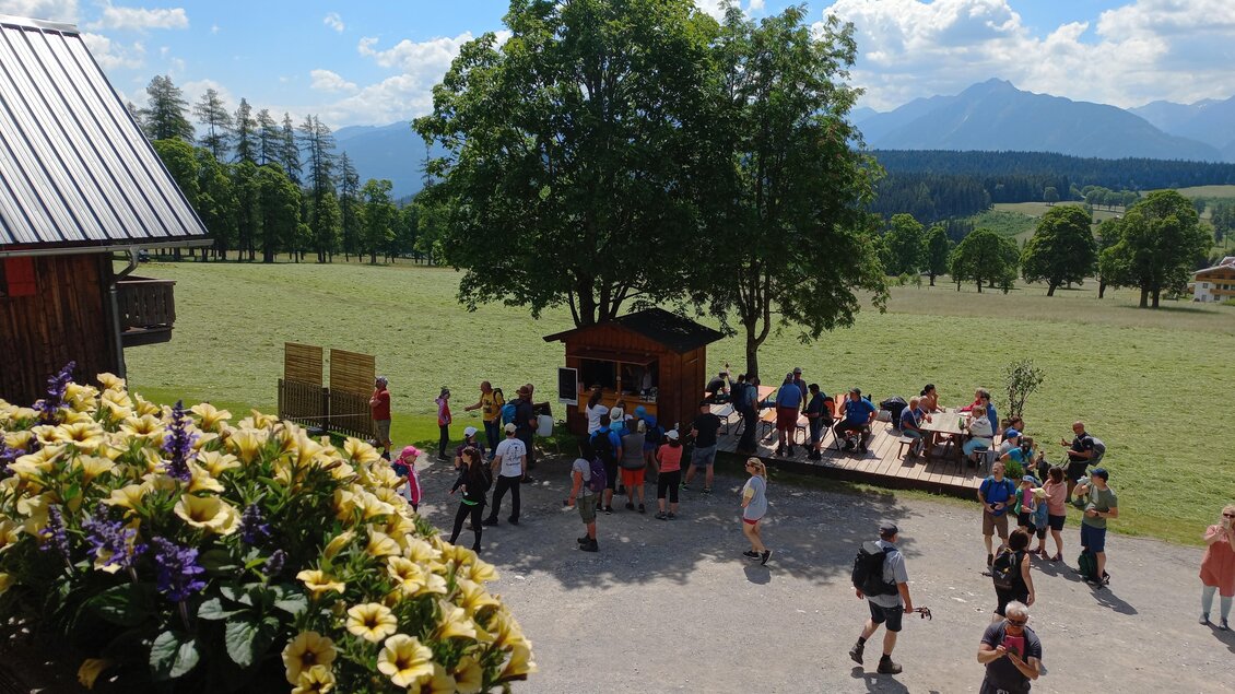 Ein belebter Außenbereich mit Menschen, die sich um eine kleine Hütte versammeln. Im Hintergrund sind Bäume und Berge unter einem blauen Himmel zu sehen. | © Tourismusverband Schladming-Dachstein