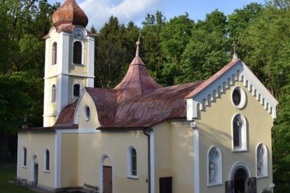 Eine kleine Kirche mit einem markanten, kupfernen Dach und einem hohen Turm. Sie steht von grünen Bäumen umgeben in einer ruhigen Umgebung.
