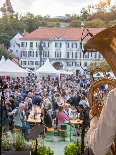 Ein lebhaftes Stadtfest mit vielen Menschen, die an Tischen sitzen und Musik genießen. Im Vordergrund spielen zwei Musiker auf der Bühne. | © TV Thermen- Vulkanland
