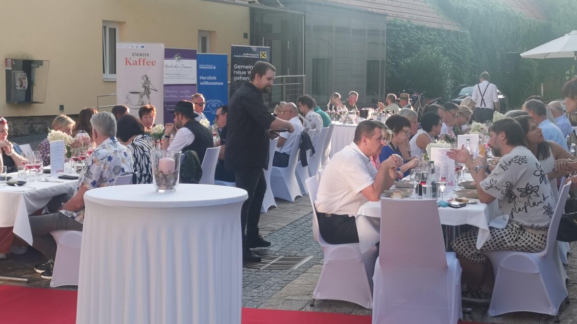 Eine festliche Veranstaltung im Freien mit vielen Gästen, die an gedeckten Tischen sitzen. Ein roter Teppich verleiht der Szenerie ein elegantes Ambiente. | © CDM incoming - Fotostudio Augenblick