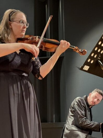 In the foreground stands Hannelore Ramminger in a black dress with her violin. She is accompanied by Philipp Scheucher at the piano, who is positioned behind her. | © Oststeiermark Tourismus