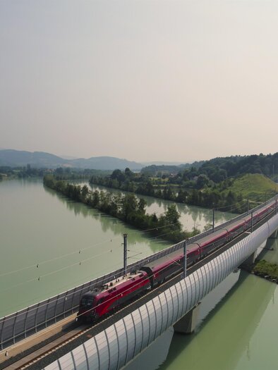 Red train of the ÖBB on the Draubrücke of the Koralmbahn, river landscape and hills in Carinthia. | © ÖBB