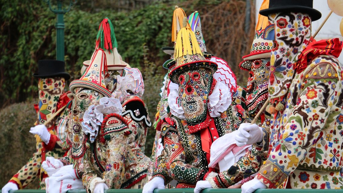 Die Ausseer Flinserl in ihren aufwendigen, bunt bestickten Kostümen und Masken stehen an einem Geländer in der Mitte von Bad Aussee. Die festliche Atmosphäre ihres traditionellen Umzugs zum Ausseer Fasching ist deutlich spürbar. | © TVB Ausseerland - Theresa Schwaiger