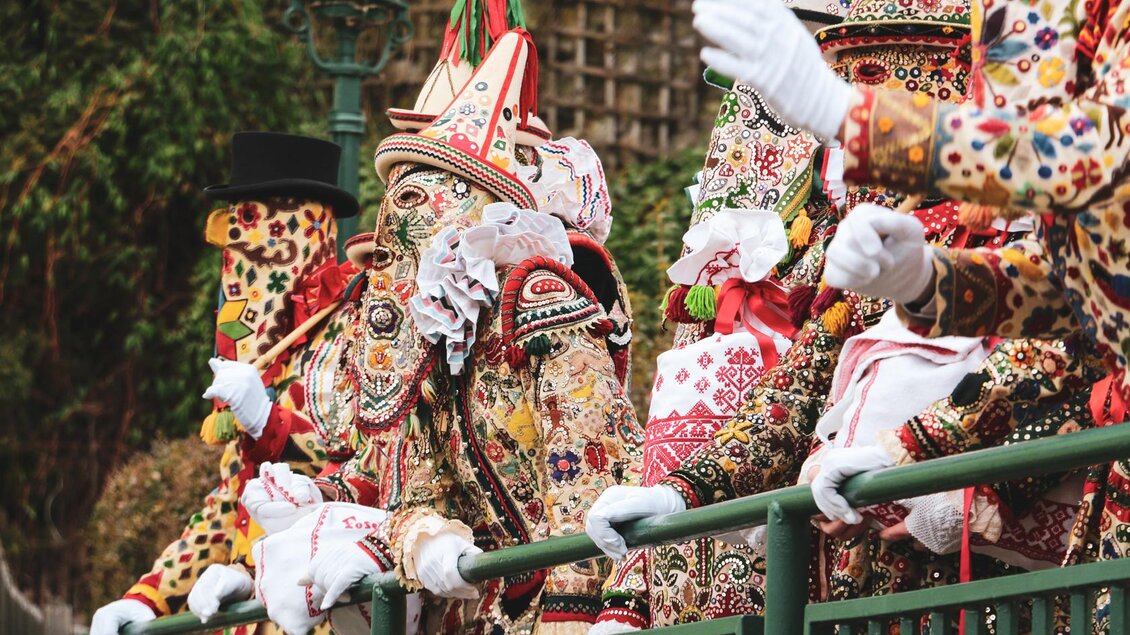Die Ausseer Flinserl in ihren aufwendigen, bunt bestickten Kostümen und Masken stehen an einem Geländer in der Mitte von Bad Aussee. Die festliche Atmosphäre ihres traditionellen Umzugs zum Ausseer Fasching ist deutlich spürbar. Die Verkleidungen sind reich verziert und zeigen verschiedene Muster. | © TVB Ausseerland - Theresa Schwaiger