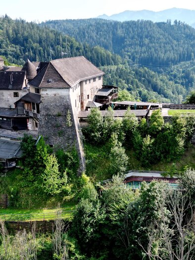 Eine alte Burg liegt malerisch auf einem Hügel, umgeben von üppigen Wäldern. Im Hintergrund erstrecken sich sanfte Berge unter einem klaren Himmel. | © Wolfgang Köllner