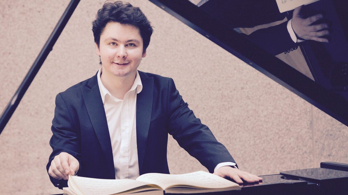 Ein junger Pianist sitzt am Klavier und blickt lächelnd in die Kamera. Vor ihm liegt ein aufgeschlagenes Notenbuch. | © A. Bashynsky