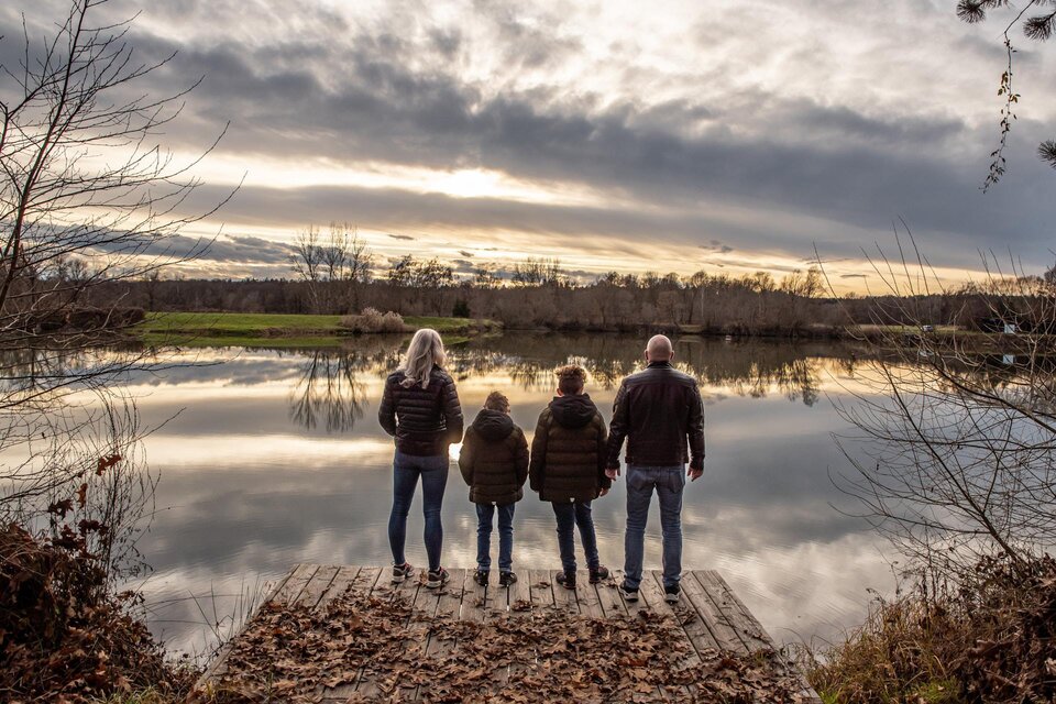 Familie steht auf Holzsteg am See, Blick über ruhiges Wasser und herbstliche Landschaft im Abendlicht. | © STMK-Michaela Brueckler