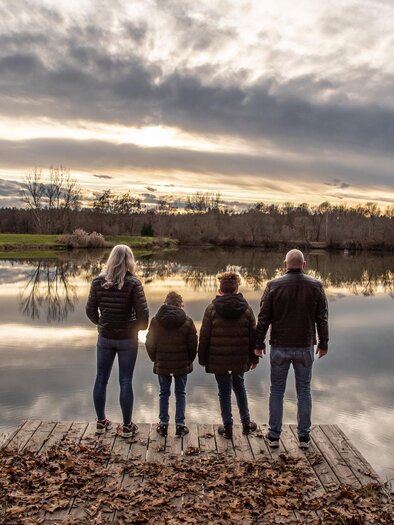 Familie steht auf Holzsteg am See, Blick über ruhiges Wasser und herbstliche Landschaft im Abendlicht. | © STMK-Michaela Brueckler