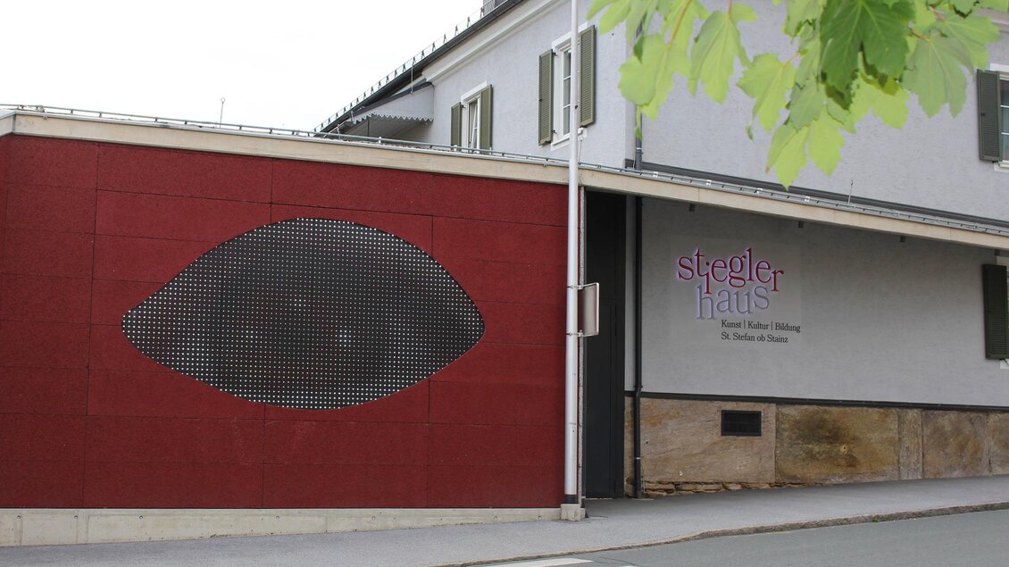 Ein modernes Gebäude mit einem auffälligen roten Wandabschnitt und einem großen, stilisierten Auge aus Lochblech. Das Schild "Stegler Haus" ist neben dem Eingang sichtbar. | © TV Südsteiermark - Irene Löschnig