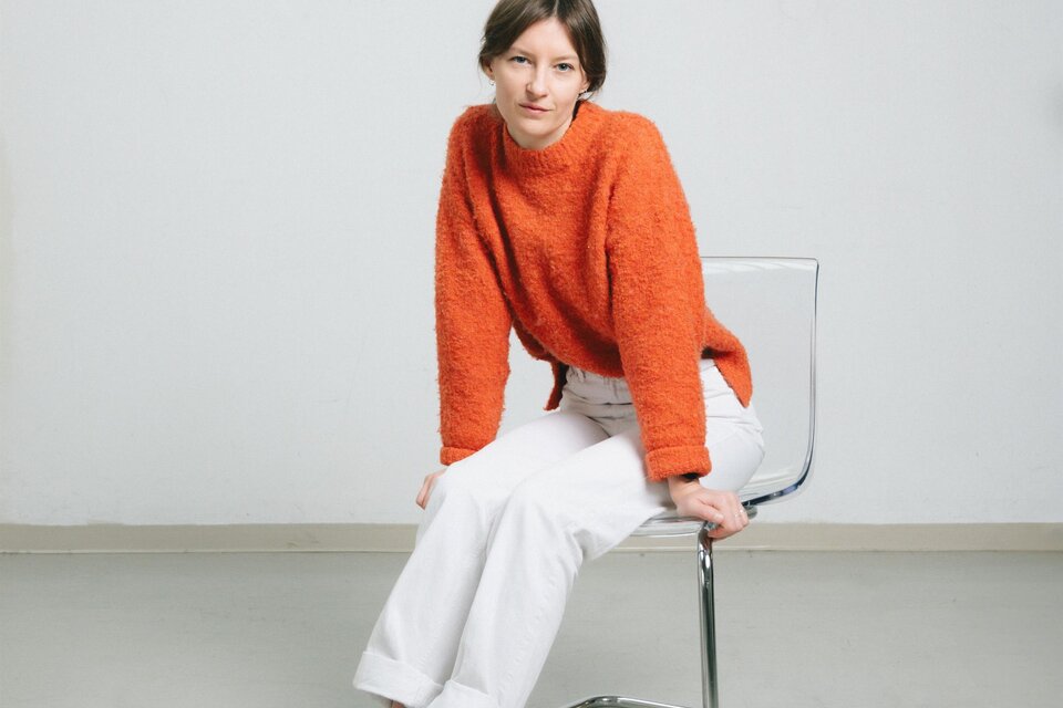 Eine Frau sitzt auf einem Stuhl und trägt einen orangefarbenen Pullover und weiße Hosen. Die Umgebung ist hell und minimalistischer Stil. | © Theresa Stritzinger