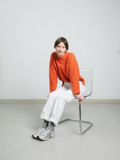Eine Frau sitzt auf einem Stuhl und trägt einen orangefarbenen Pullover und weiße Hosen. Die Umgebung ist hell und minimalistischer Stil. | © Theresa Stritzinger