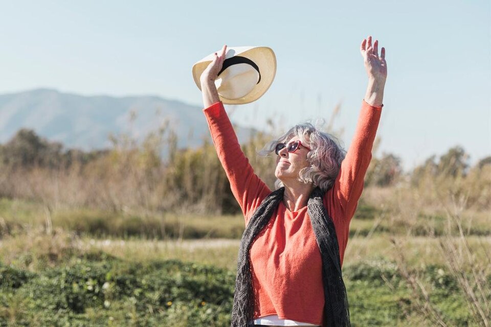 Eine Frau mit grauen Haaren hebt ihre Arme in die Luft und trägt einen Hut. Sie strahlt Freude und Zufriedenheit aus vor einer grünen Landschaft. | © Bild: freepik