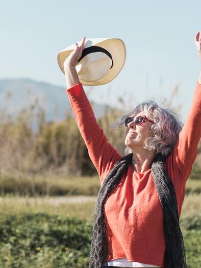 A cheerful older woman stands outdoors, raising her hands in the air. She is wearing a hat and sunglasses, and in the background, gentle hills can be seen. | © Bild: freepik